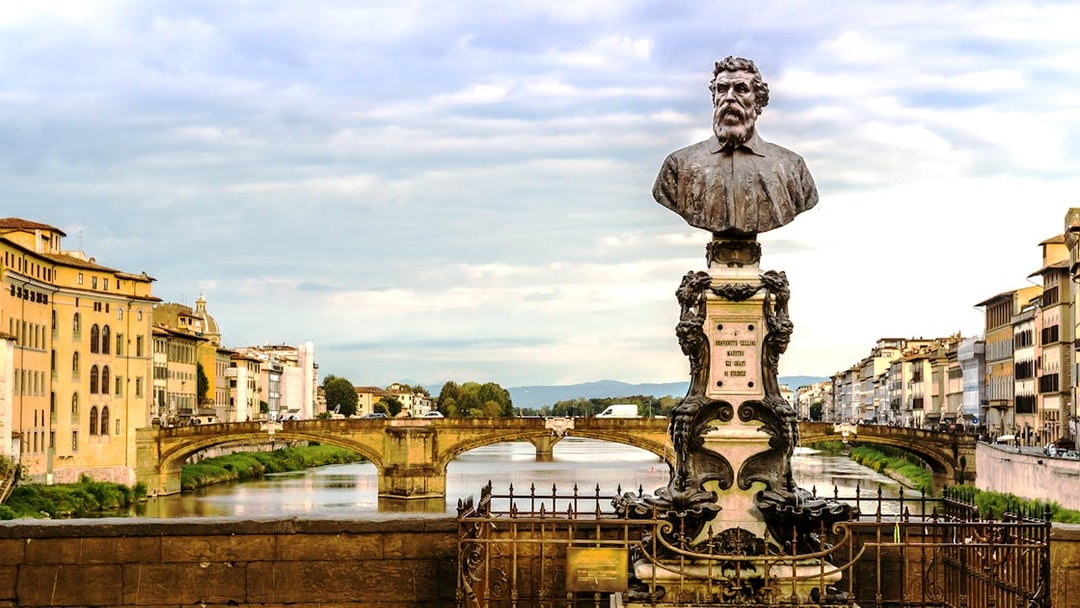 Bust of Benvenuto Cellini on Ponte Vecchio, Florence, with Arno River and historic buildings in view.