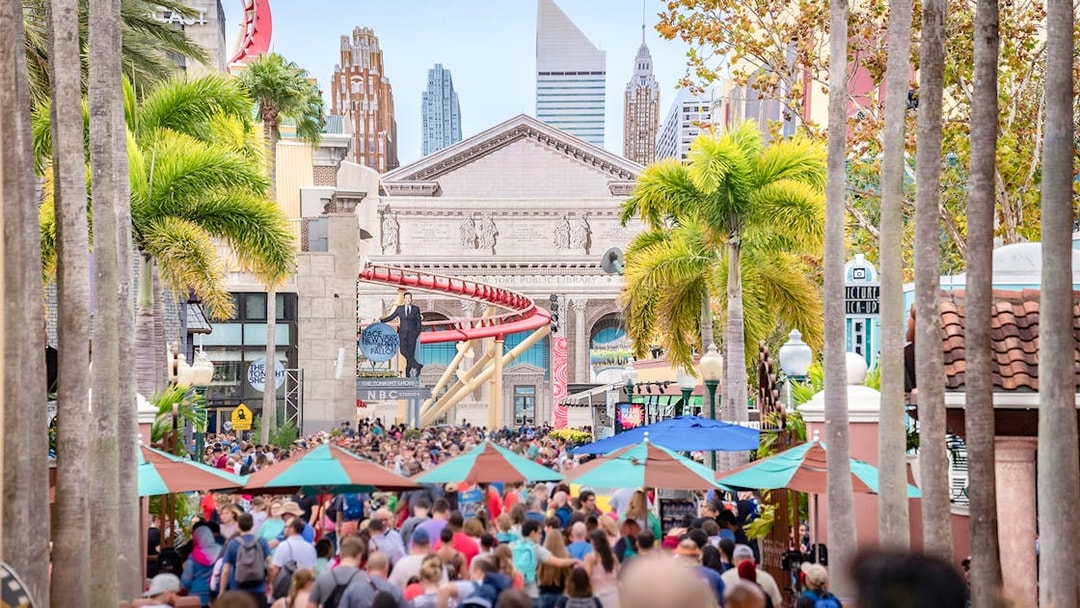 Crowd entering Universal Studios Resort, Orlando, with palm trees and cityscape backdrop.