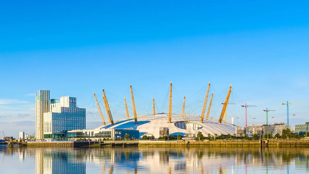 O2 Arena in London with surrounding buildings and cranes reflected in the Thames River.