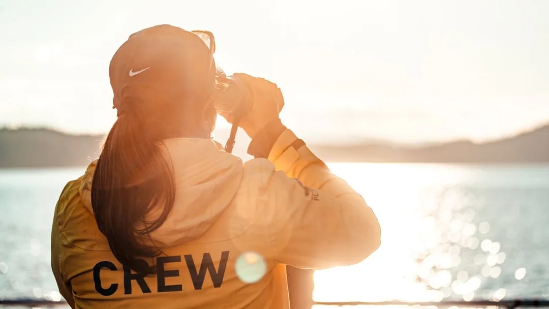 Person in yellow jacket with "CREW" on back, looking through binoculars over a sunlit lake.