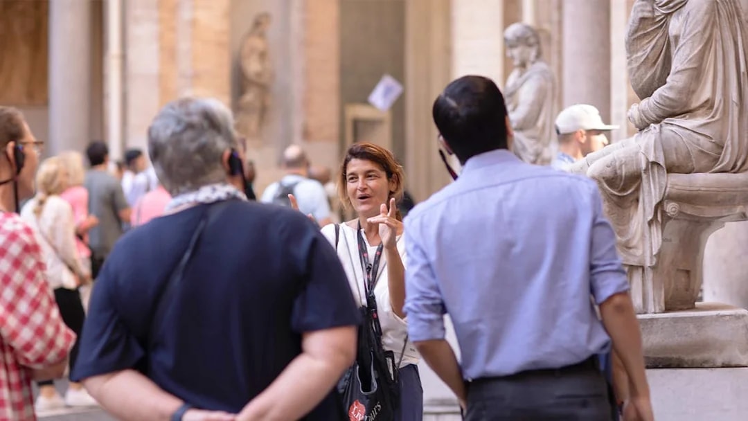 Guide leading a small tour group at the Vatican Museum, standing near classical statues.