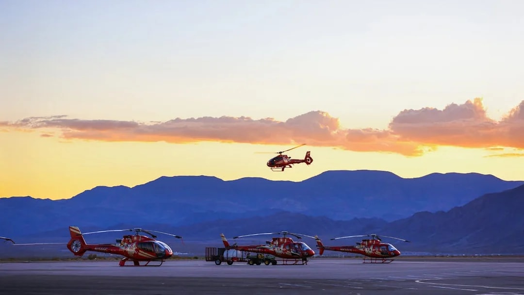 Helicopters parked on tarmac with one in flight at sunset, mountains in background.
