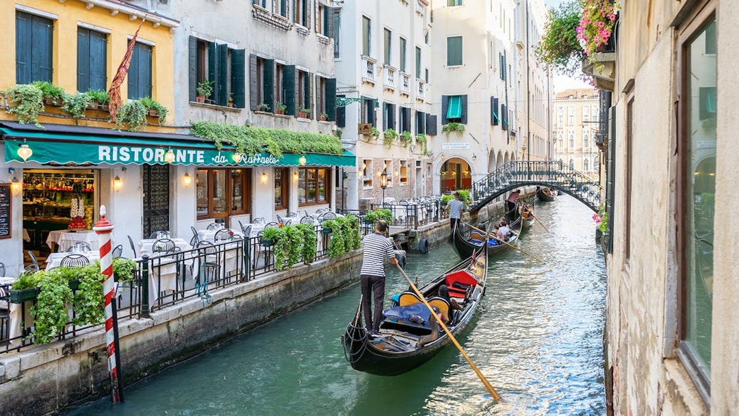 Gondolas navigating a canal in Venice near a restaurant, with a bridge in the background.