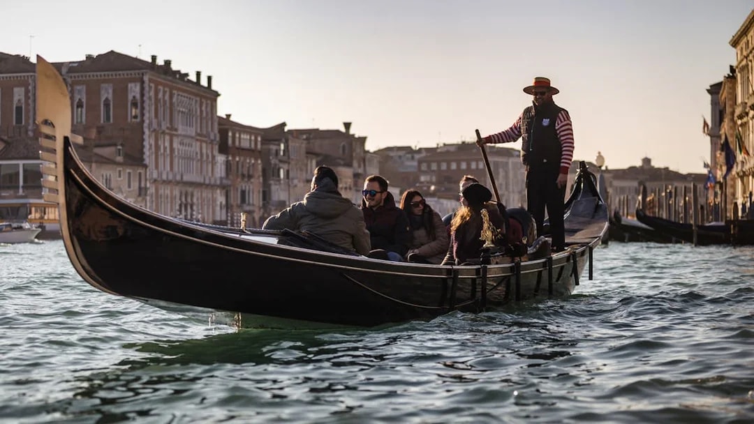 Gondola ride on Venice canal with tourists and gondolier, historic buildings in background.