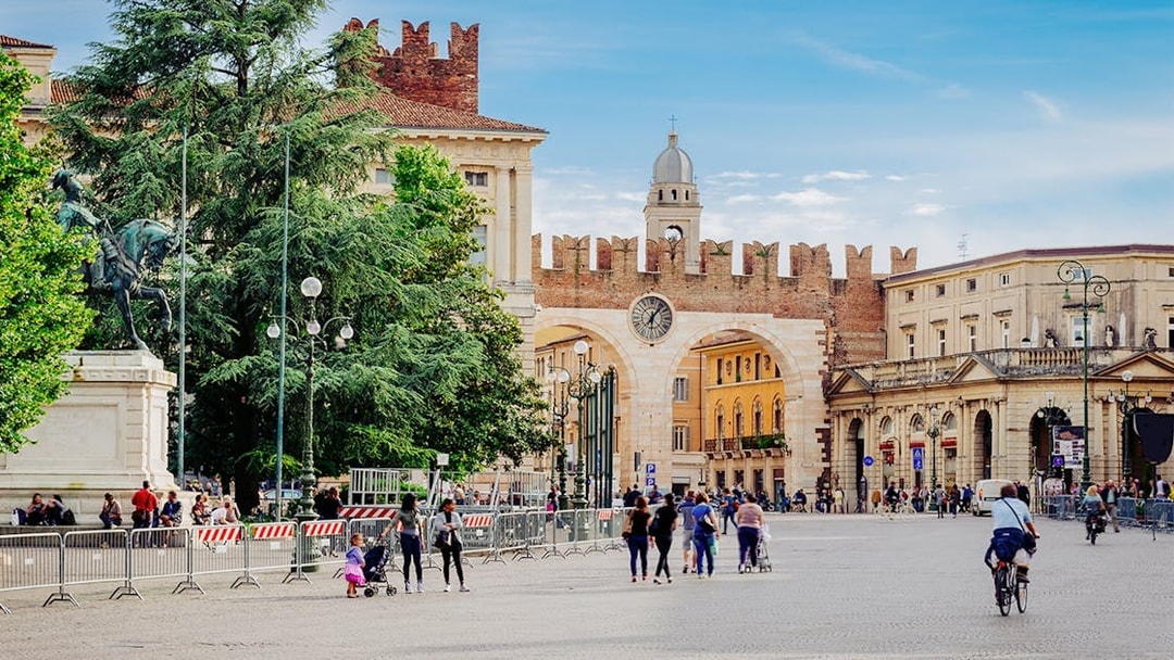 Medieval gates and clock tower on Piazza Bra, Verona, Italy, with people walking nearby.