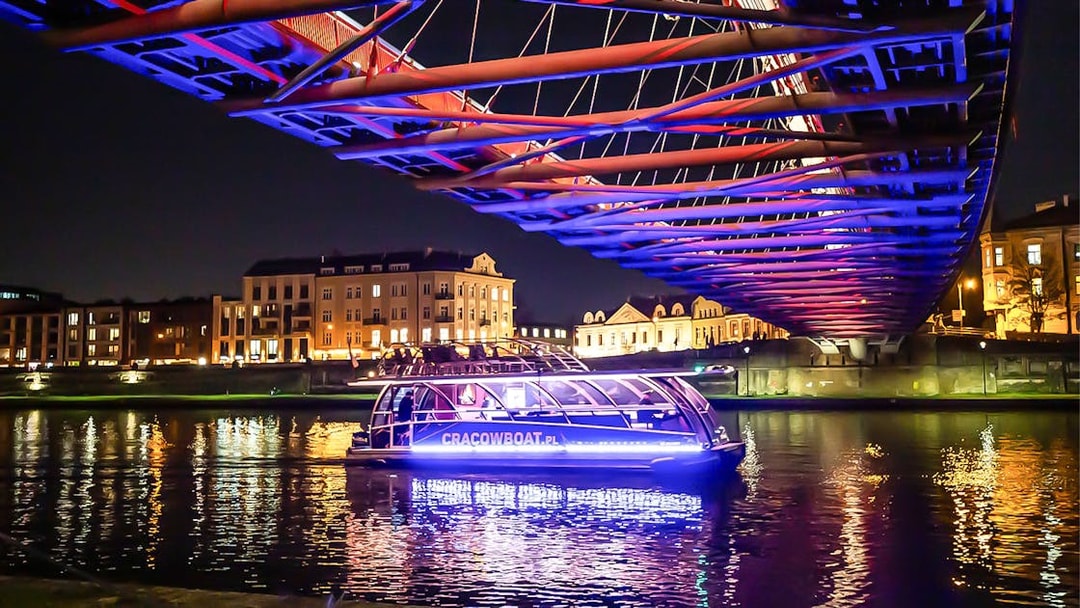 Catamaran on Vistula River at night under illuminated bridge in Krakow.