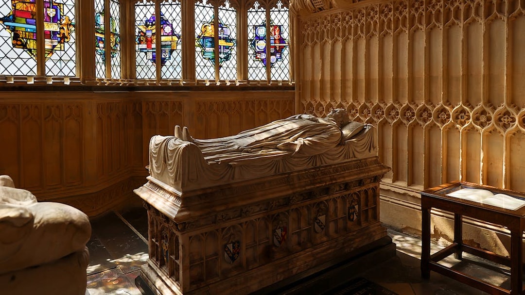 Tomb with effigy inside Westminster Abbey, London, with stained glass windows in the background.