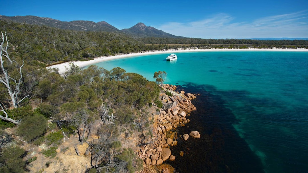 Cruise boat in turquoise waters of Wineglass Bay, Tasmania, with forested hills and rocky shoreline.