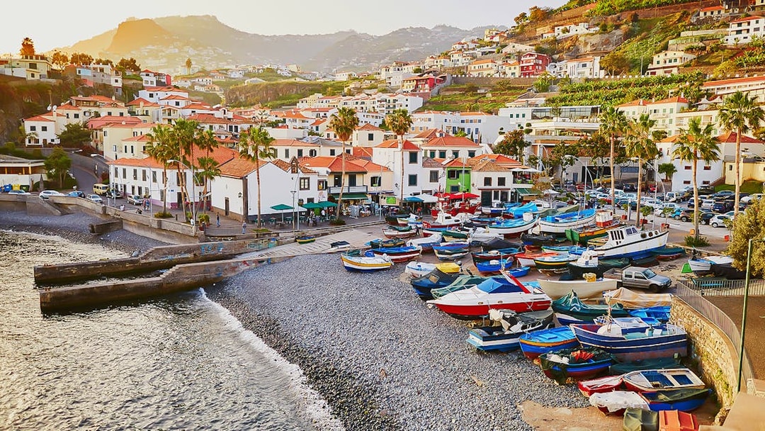 Colorful boats on a pebble beach in Madeira with hillside houses in the background.