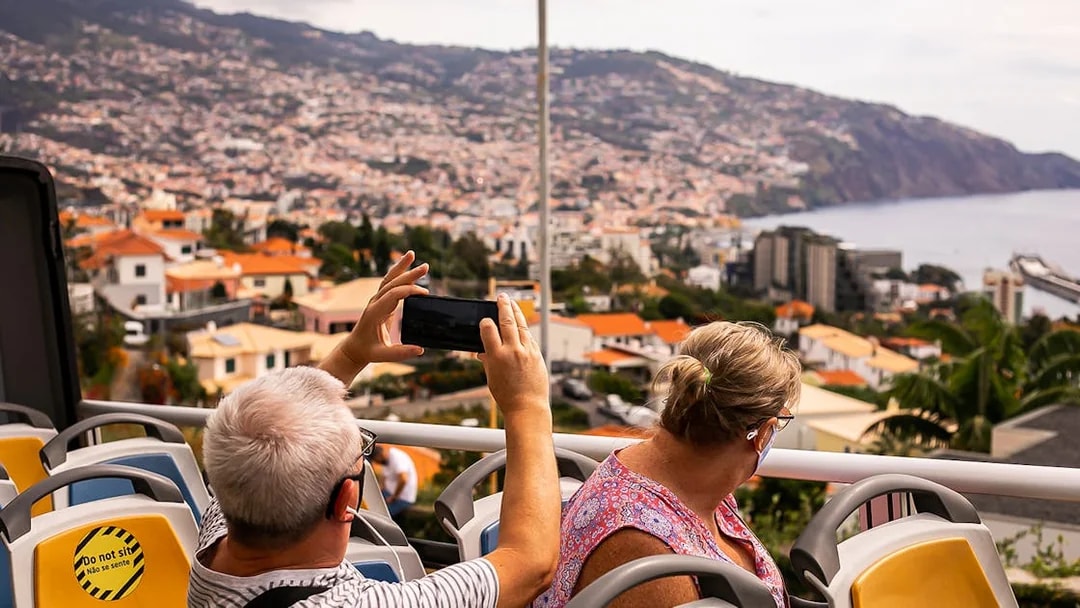 Tourists on Madeira hop-on hop-off bus capturing cityscape views.