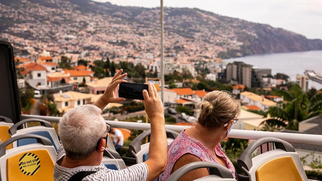 Tourists on a Funchal bus tour capturing city views with mountains and ocean in the background.