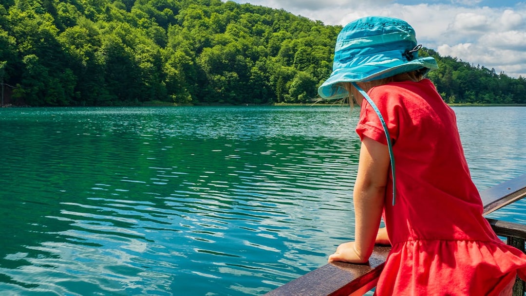 Child in blue hat looking at Plitvice Lakes from a boat, surrounded by lush greenery.