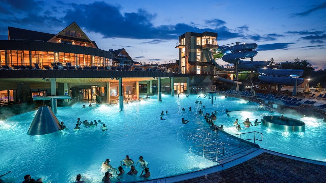 Tourists relaxing in Chochołow Thermal Baths with mountain view, evening lights, Zakopane transfers available.