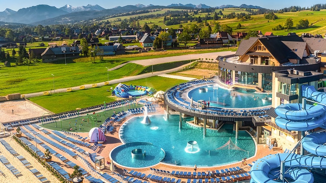 Tourists relaxing in a thermal spa with pools and slides in Zakopane, Poland, surrounded by scenic mountains.